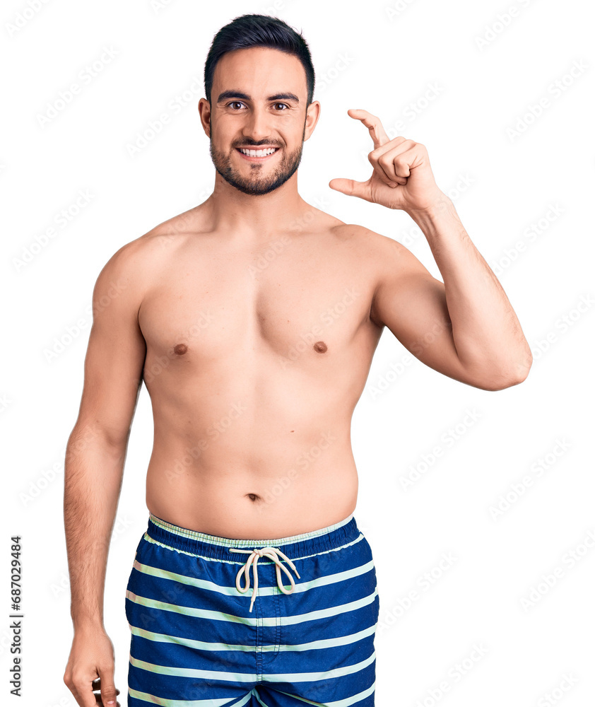 Young handsome man wearing swimwear smiling and confident gesturing with hand doing small size sign with fingers looking and the camera. measure concept. Young handsome man wearing swimwear smiling and confident gesturing with hand doing small size sign with fingers looking and the camera. measure concept.