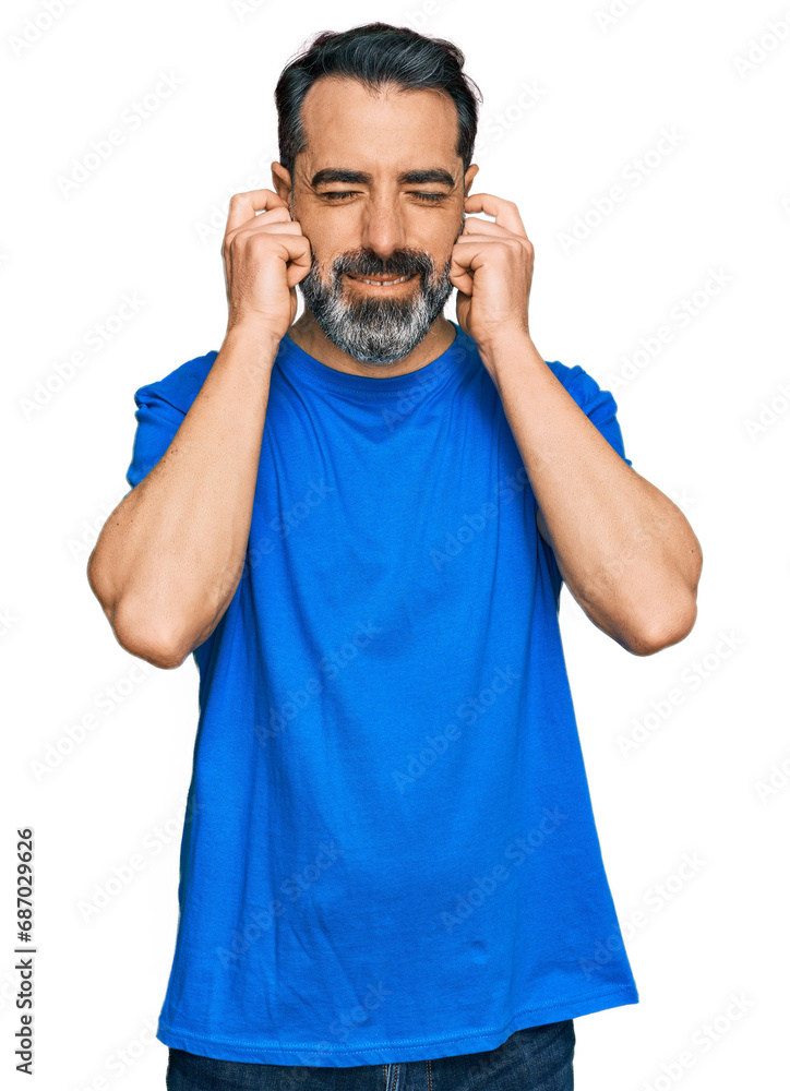 Middle aged man with beard wearing casual blue t shirt covering ears with fingers with annoyed expression for the noise of loud music. deaf concept. Middle aged man with beard wearing casual blue t shirt covering ears with fingers with annoyed expression for the noise of loud music. deaf concept.