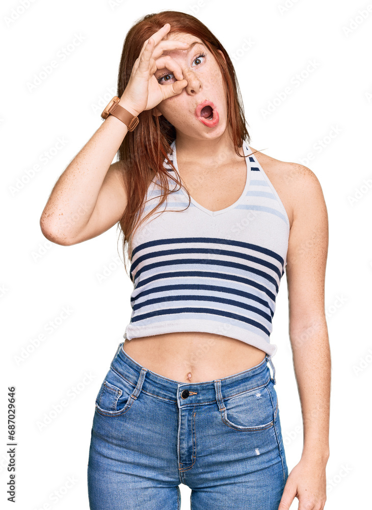 Young read head woman wearing casual clothes doing ok gesture shocked with surprised face, eye looking through fingers. unbelieving expression.