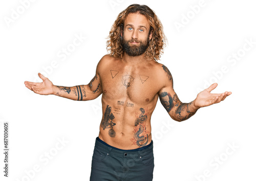 Handsome man with beard and long hair standing shirtless showing tattoos clueless and confused expression with arms and hands raised. doubt concept.