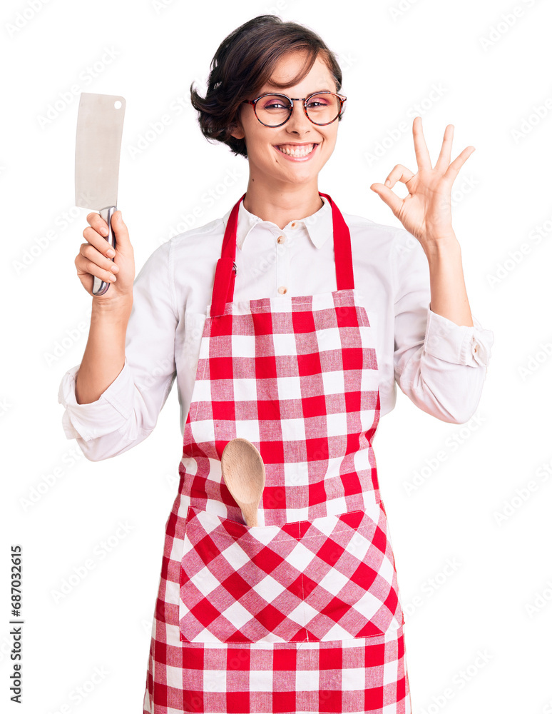 Beautiful young woman with short hair wearing professional baker apron holding knife doing ok sign with fingers, smiling friendly gesturing excellent symbol Beautiful young woman with short hair wearing professional baker apron holding knife doing ok sign with fingers, smiling friendly gesturing excellent symbol