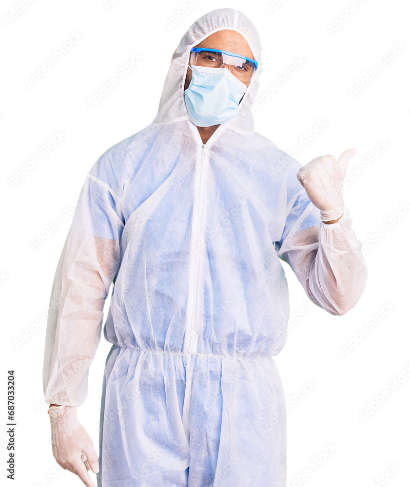 Young hispanic man wearing doctor protection coronavirus uniform and medical mask smiling with happy face looking and pointing to the side with thumb up. Young hispanic man wearing doctor protection coronavirus uniform and medical mask smiling with happy face looking and pointing to the side with thumb up.
