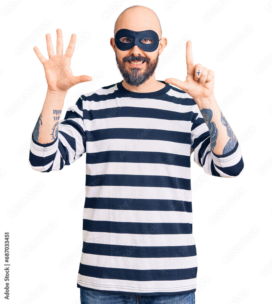Young handsome man wearing burglar mask showing and pointing up with fingers number seven while smiling confident and happy. Young handsome man wearing burglar mask showing and pointing up with fingers number seven while smiling confident and happy.
