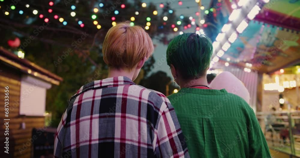 Rear view of a blonde girl with a short haircut in a plaid shirt and a girl with a green short haircut in a green shirt walk through a bright and glowing amusement park during their date Rear view of a blonde girl with a short haircut in a plaid shirt and a girl with a green short haircut in a green shirt walk through a bright and glowing amusement park during their date