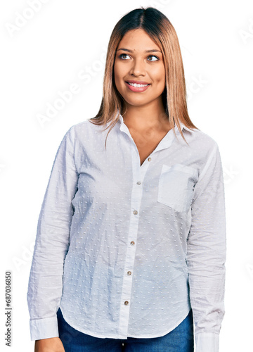 Young hispanic woman wearing casual white shirt looking away to side with smile on face, natural expression. laughing confident.