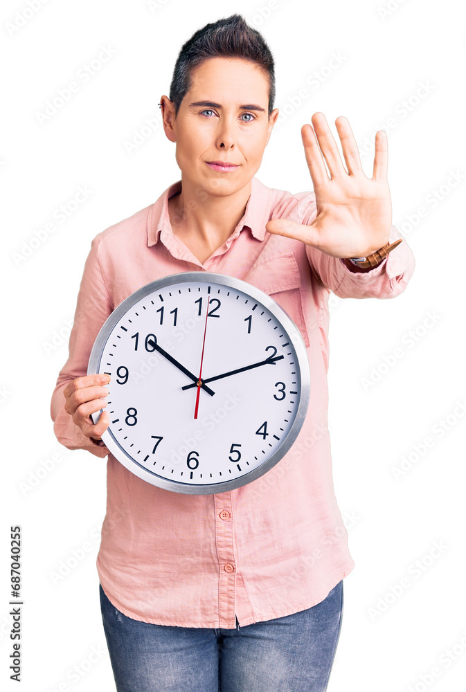 Young woman with short hair holding big clock with open hand doing stop sign with serious and confident expression, defense gesture Young woman with short hair holding big clock with open hand doing stop sign with serious and confident expression, defense gesture