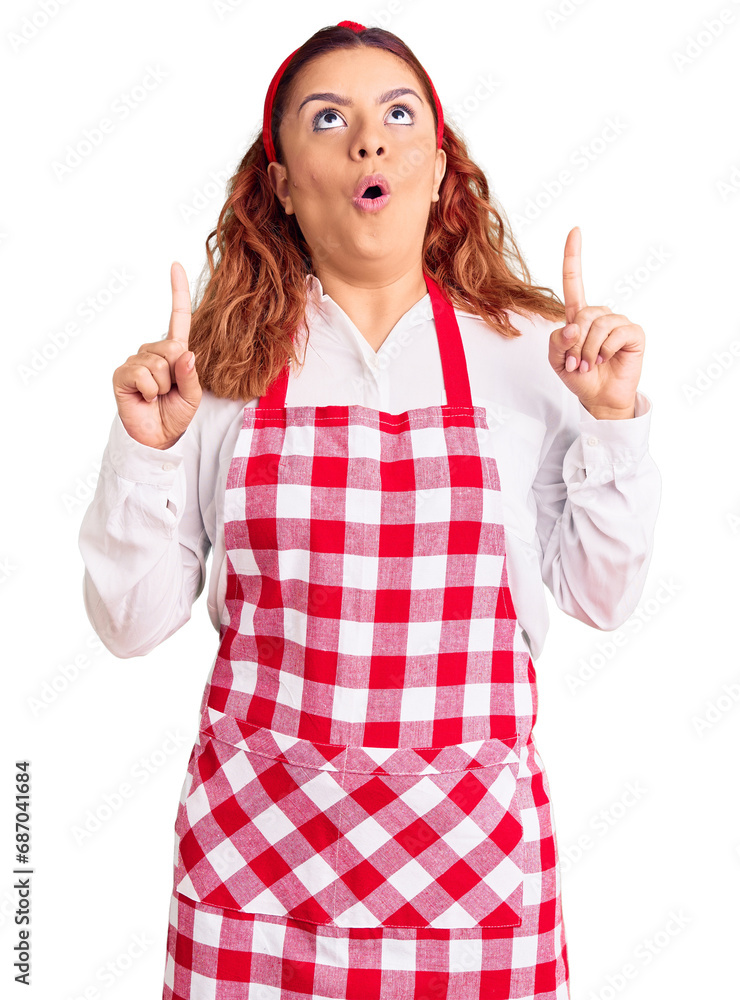 Young latin woman wearing apron amazed and surprised looking up and pointing with fingers and raised arms. Young latin woman wearing apron amazed and surprised looking up and pointing with fingers and raised arms.