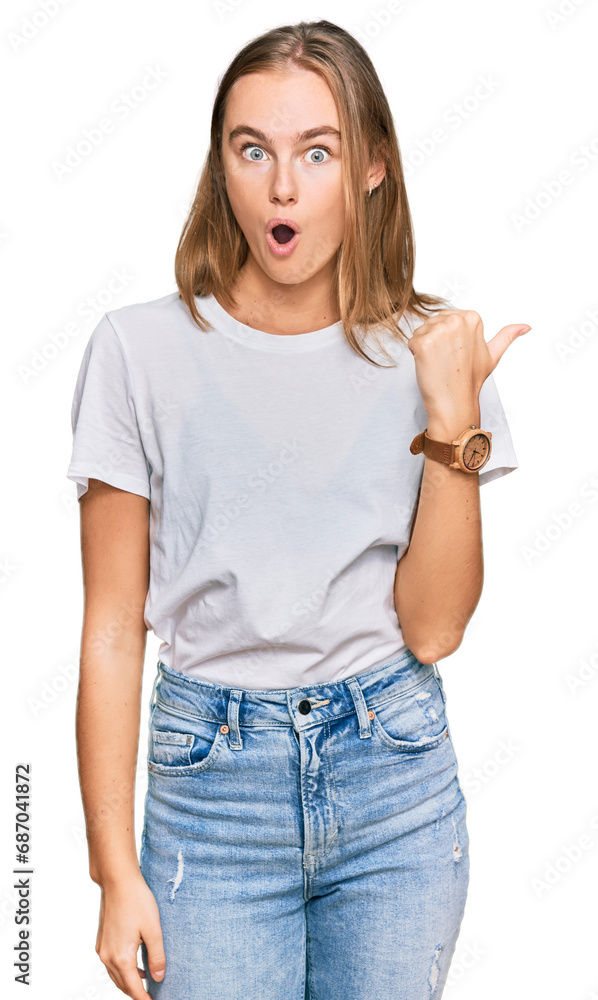 Obraz premium Beautiful young blonde woman wearing casual white t shirt surprised pointing with hand finger to the side, open mouth amazed expression.