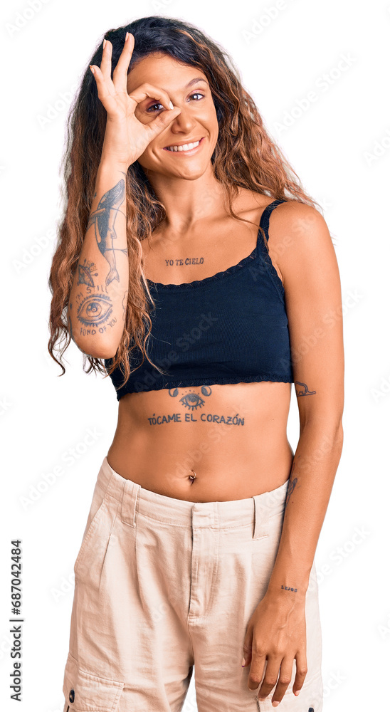 Young hispanic woman with tattoo wearing casual clothes doing ok gesture with hand smiling, eye looking through fingers with happy face.