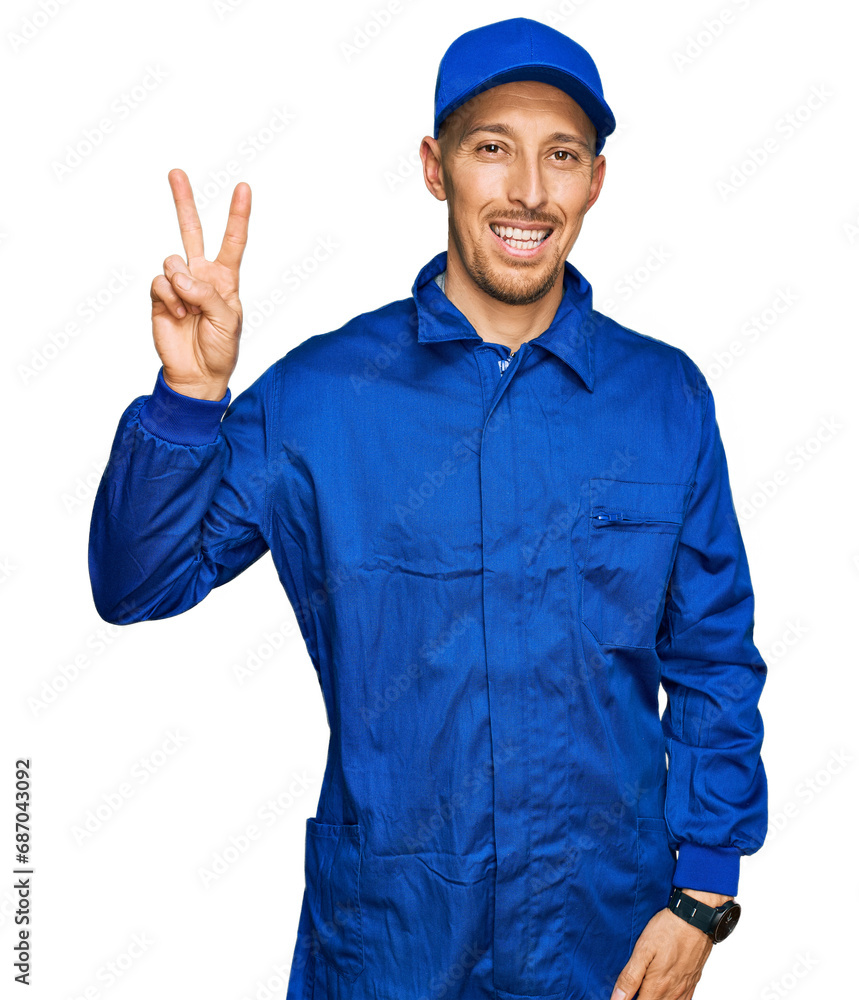 Bald man with beard wearing builder jumpsuit uniform smiling looking to the camera showing fingers doing victory sign. number two. Bald man with beard wearing builder jumpsuit uniform smiling looking to the camera showing fingers doing victory sign. number two.