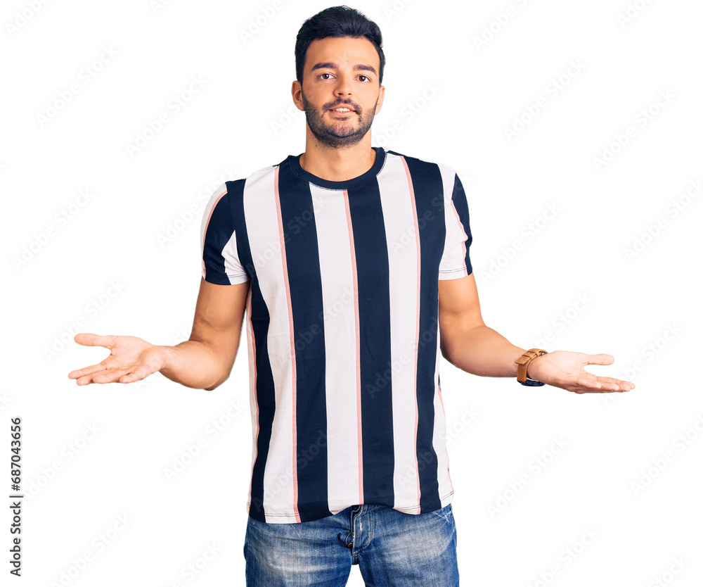 Naklejka premium Young handsome hispanic man wearing striped tshirt clueless and confused expression with arms and hands raised. doubt concept.