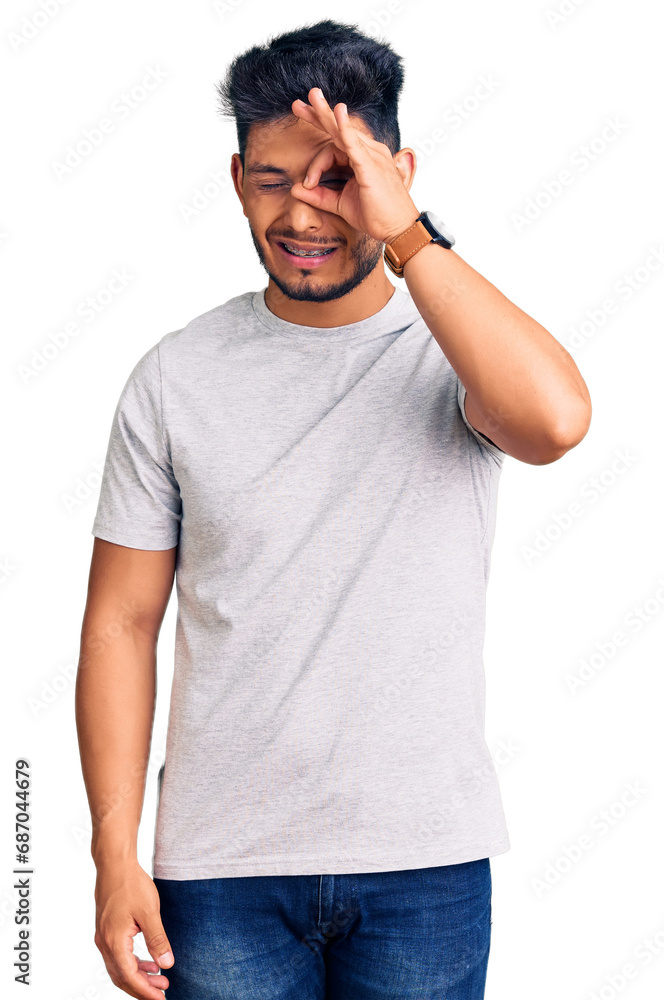 Handsome latin american young man wearing casual tshirt doing ok gesture with hand smiling, eye looking through fingers with happy face. Handsome latin american young man wearing casual tshirt doing ok gesture with hand smiling, eye looking through fingers with happy face.