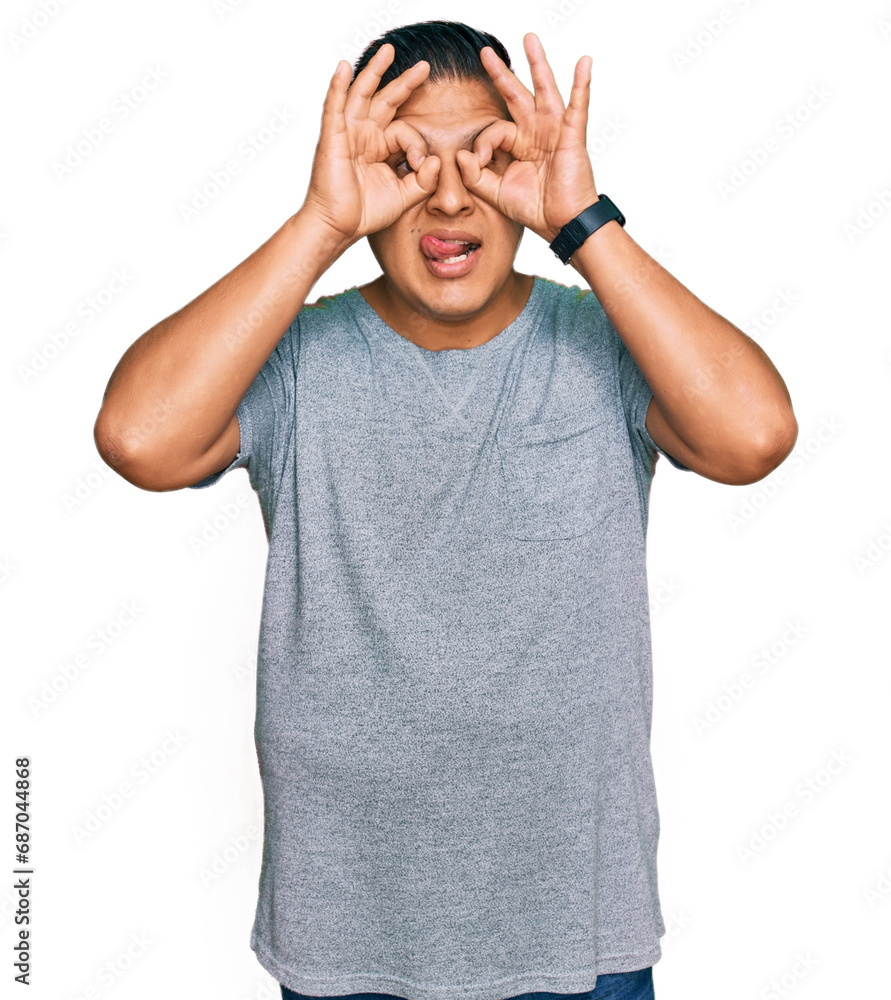 Fototapeta premium Young latin man wearing casual clothes doing ok gesture like binoculars sticking tongue out, eyes looking through fingers. crazy expression.