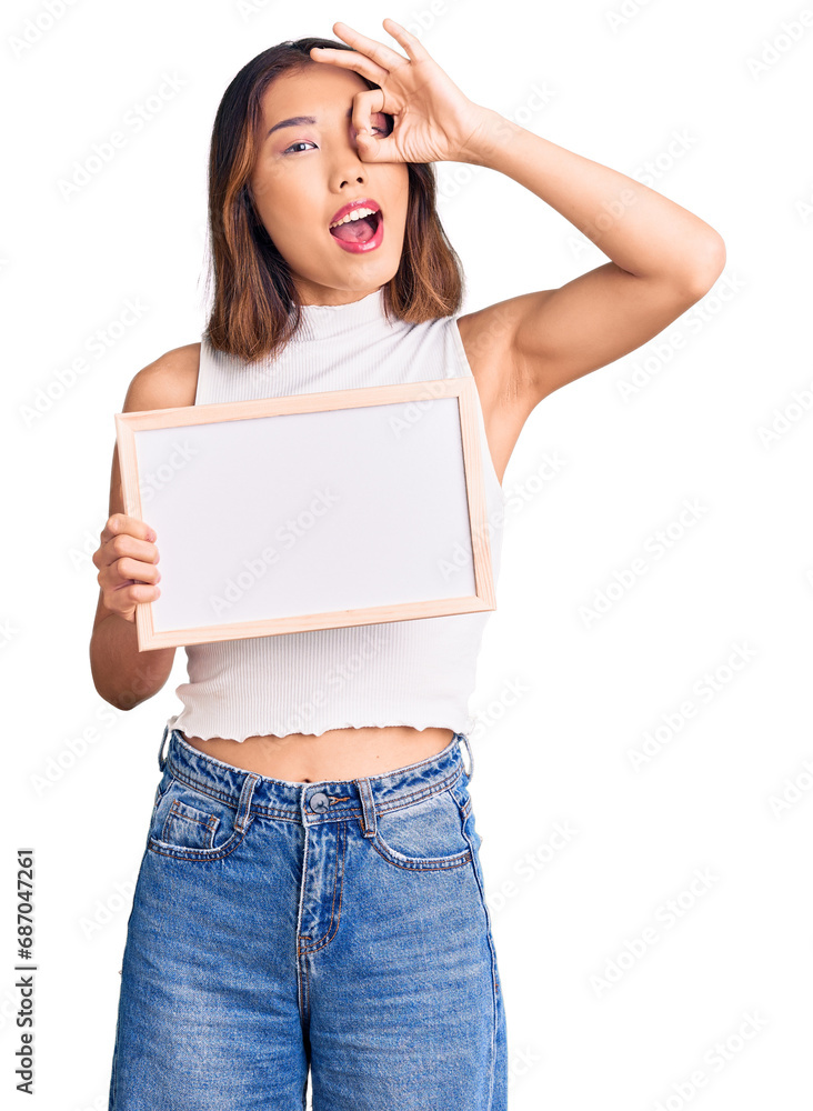 Young beautiful chinese girl holding empty white chalkboard smiling happy doing ok sign with hand on eye looking through fingers Young beautiful chinese girl holding empty white chalkboard smiling happy doing ok sign with hand on eye looking through fingers