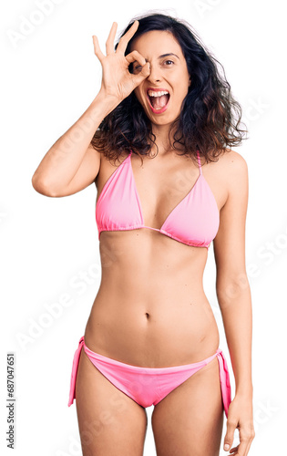 Young beautiful hispanic woman wearing bikini doing ok gesture with hand smiling, eye looking through fingers with happy face.