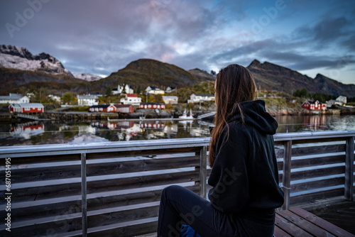 Obraz na płótnie young woman drinking coffee in the morning and taking a look on the bay in Sørvågen fisherman village with red houses on the coast, lofoten islands, norway w salonie