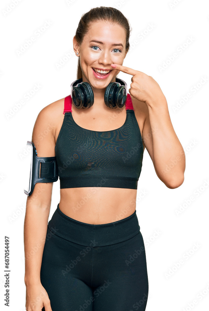Beautiful blonde woman wearing gym clothes and using headphones pointing with hand finger to face and nose, smiling cheerful. beauty concept Beautiful blonde woman wearing gym clothes and using headphones pointing with hand finger to face and nose, smiling cheerful. beauty concept