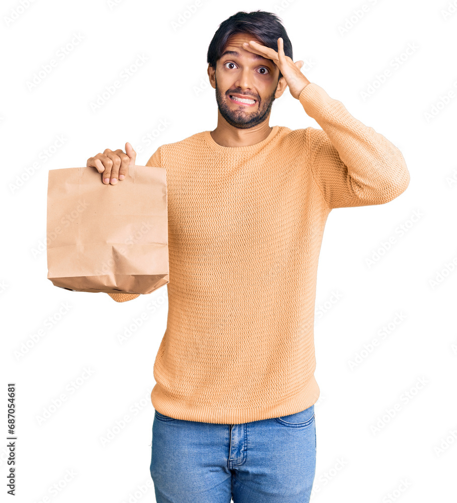 Fototapeta premium Handsome hispanic man holding take away paper bag stressed and frustrated with hand on head, surprised and angry face