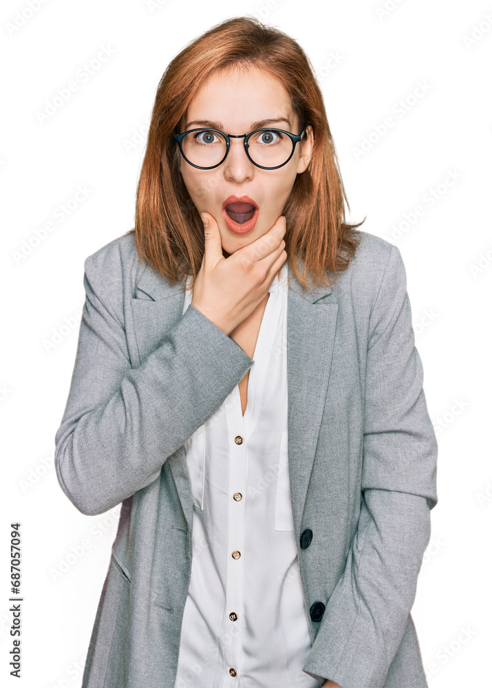 Young caucasian woman wearing business style and glasses looking fascinated with disbelief, surprise and amazed expression with hands on chin Young caucasian woman wearing business style and glasses looking fascinated with disbelief, surprise and amazed expression with hands on chin