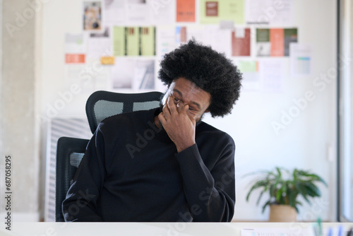 Black businessman with afro look sitting at his desk in the office. He rubs his eyes because he is tired. Overtime at work photo