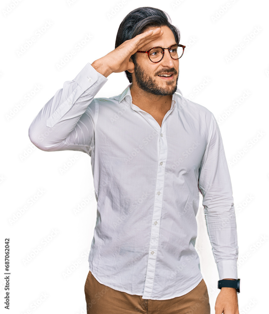 Young hispanic man wearing business shirt and glasses very happy and smiling looking far away with hand over head. searching concept. Young hispanic man wearing business shirt and glasses very happy and smiling looking far away with hand over head. searching concept.