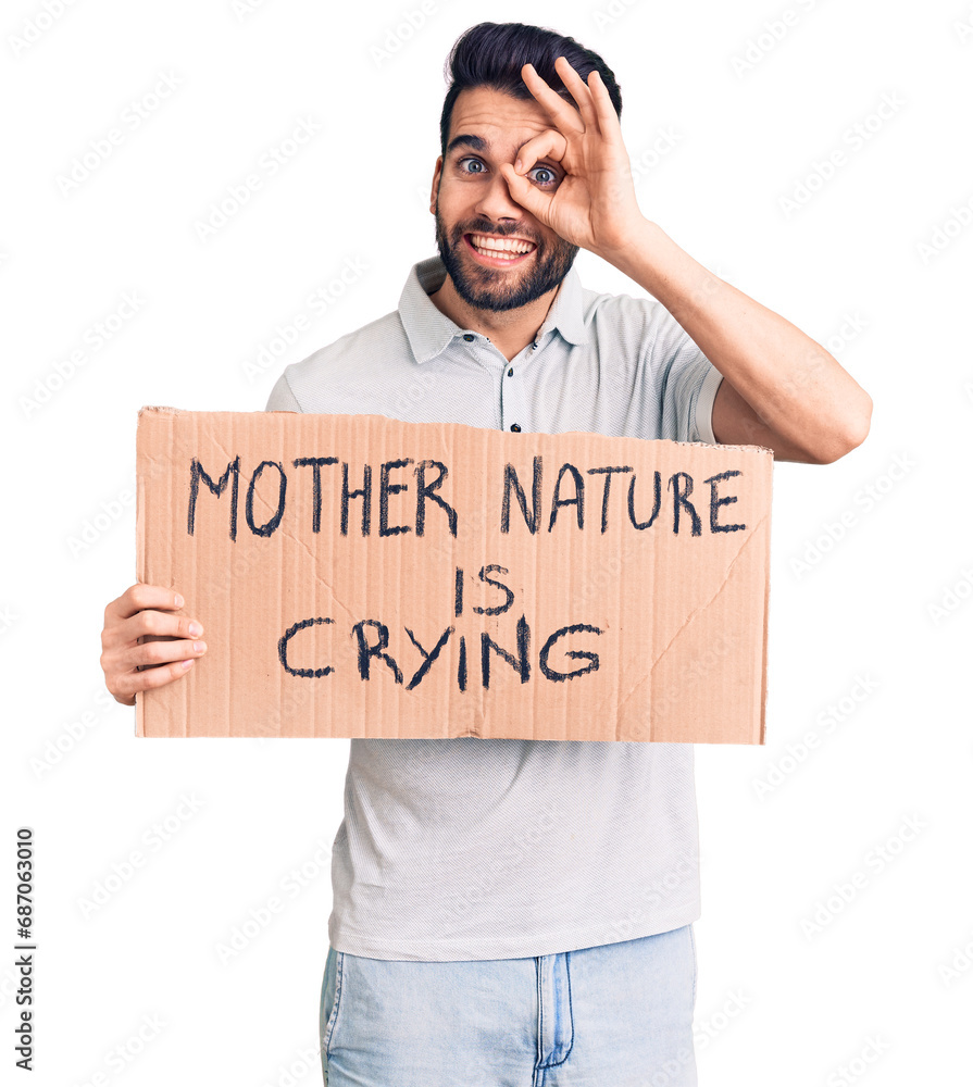 Young handsome man with beard holding mother nature is crying cardboard banner smiling happy doing ok sign with hand on eye looking through fingers Young handsome man with beard holding mother nature is crying cardboard banner smiling happy doing ok sign with hand on eye looking through fingers