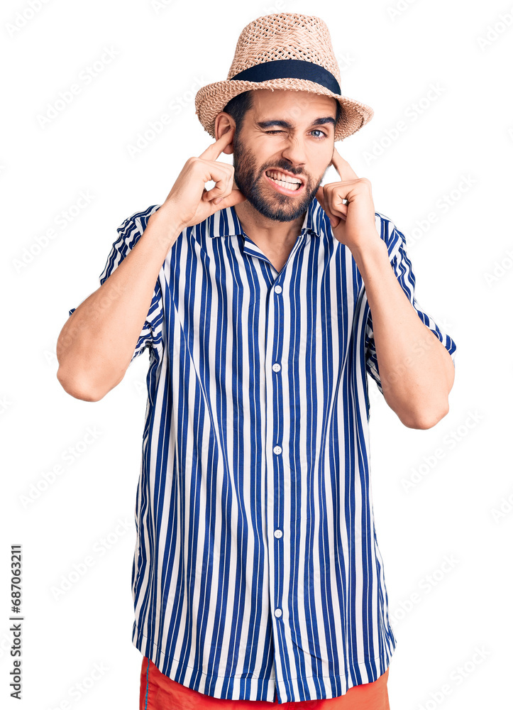 Young handsome man with beard wearing summer hat and striped shirt covering ears with fingers with annoyed expression for the noise of loud music. deaf concept. Young handsome man with beard wearing summer hat and striped shirt covering ears with fingers with annoyed expression for the noise of loud music. deaf concept.