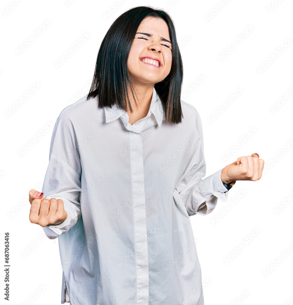 Young brunette woman with blue eyes wearing oversize white shirt very happy and excited doing winner gesture with arms raised, smiling and screaming for success. celebration concept. Young brunette woman with blue eyes wearing oversize white shirt very happy and excited doing winner gesture with arms raised, smiling and screaming for success. celebration concept.