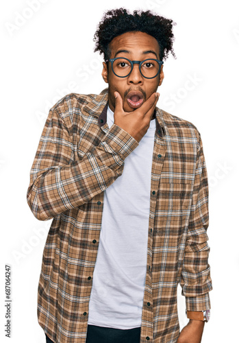 Young african american man with beard wearing casual clothes and glasses looking fascinated with disbelief, surprise and amazed expression with hands on chin