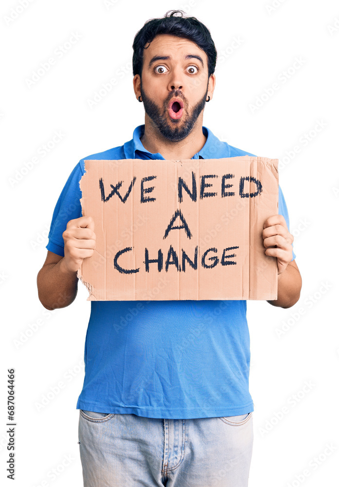 Young hispanic man holding we need a change banner scared and amazed with open mouth for surprise, disbelief face Young hispanic man holding we need a change banner scared and amazed with open mouth for surprise, disbelief face