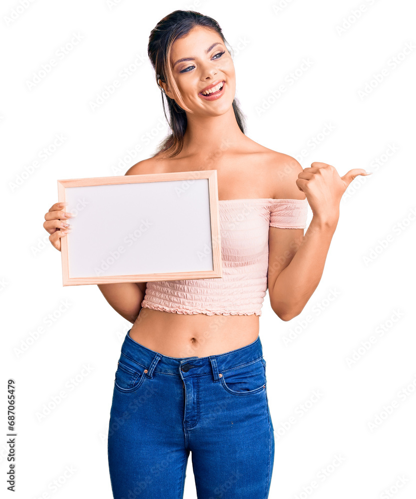 Young beautiful woman holding empty white chalkboard pointing thumb up to the side smiling happy with open mouth Young beautiful woman holding empty white chalkboard pointing thumb up to the side smiling happy with open mouth