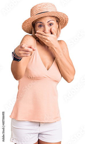 Young blonde woman wearing summer hat laughing at you, pointing finger to the camera with hand over mouth, shame expression