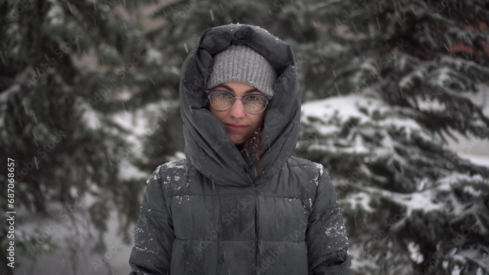 A young woman stands against a background of fir trees under heavy snowfall in winter. A girl in glasses and a down jacket with a hood stands and looks at the camera. Closeup. A young woman stands against a background of fir trees under heavy snowfall in winter. A girl in glasses and a down jacket with a hood stands and looks at the camera. Closeup.