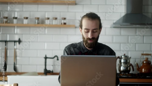 Bearded young adult freelancer man sitting in the kitchen working with laptop, getting works done photo