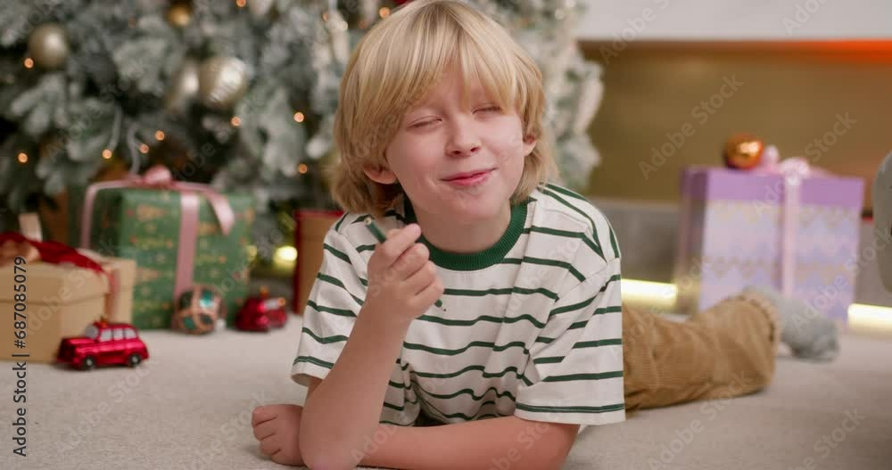 blonde boy writes a letter with wish list to santa claus using color pencils laying on floor near christmas tree. blonde boy writes a letter with wish list to santa claus using color pencils laying on floor near christmas tree.