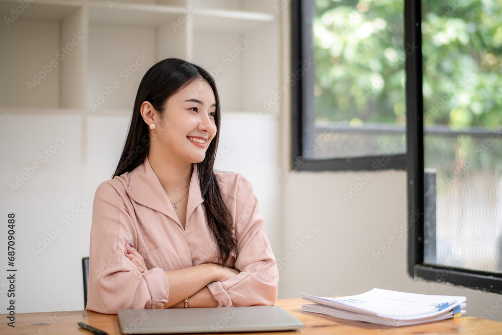 Asian businesswoman sits with her arms crossed at a desk with documents on the table. Asian businesswoman sits with her arms crossed at a desk with documents on the table.