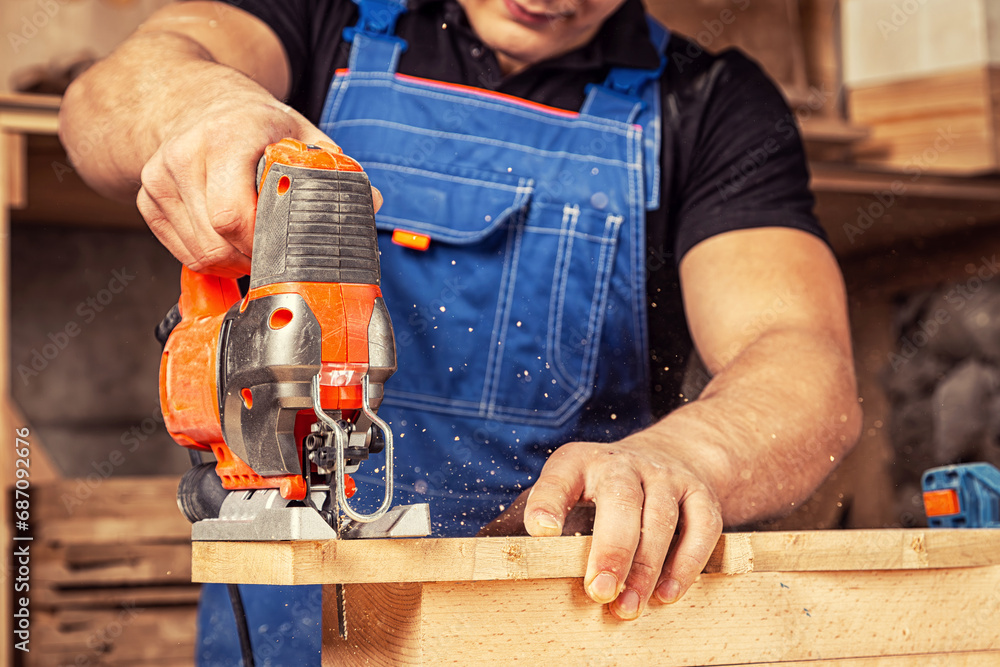 Close up of experienced carpenter in work clothes and small buiness owner carpenter saw and processes the edges of a wooden bar with a jig saw in a workshop Close up of experienced carpenter in work clothes and small buiness owner carpenter saw and processes the edges of a wooden bar with a jig saw in a workshop