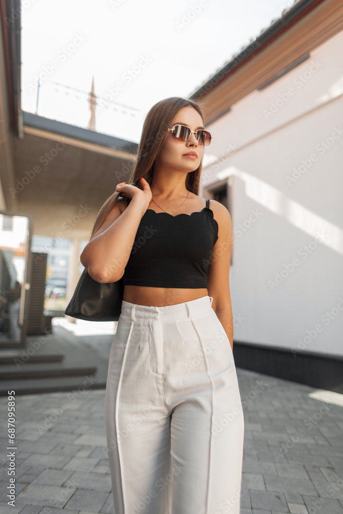 Beautiful fresh beauty stylish model girl with sunglasses in fashionable clothes with top, white pants and bag walking on the street