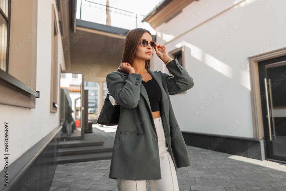 Beautiful fashion business girl in fashionable clothes with a stylish green blazer with a bag puts on glasses and stands in the city near the building Beautiful fashion business girl in fashionable clothes with a stylish green blazer with a bag puts on glasses and stands in the city near the building