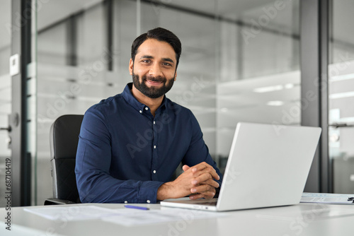 Smiling confident Indian business man employee looking at camera sitting at work desk with laptop computer. Portrait of smart happy businessman office worker or entrepreneur posing at modern workplace photo