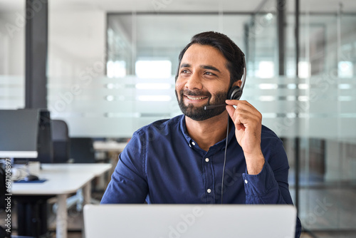 Smiling happy Indian call center agent wearing headset talking to client, contract service telemarketing operator using laptop having conversation working in customer tech assistance support office. photo