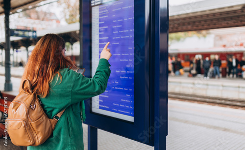 Young redhead woman with backpack pointing finger on train timetable on a railway station. 30s female with smart phone Looking at Arrival and Departure Information Display, Traveler