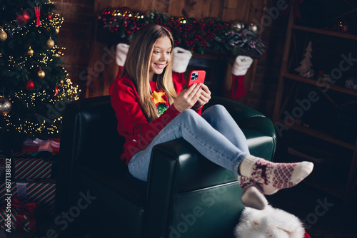 Full size photo of clever cheerful teenager with blond hair dressed red sweater sitting on armachir look at smartphone at home indoors