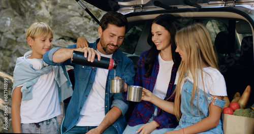 Family woman and man with teen children have picnic in nature  they have stop with car sitting in trunk and drinking tea from thermos. Family relationships  dating  companionship  road trip  traveling