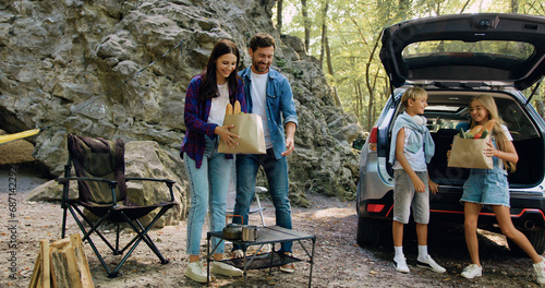 Parents and children take out unpacking packages of products from open trunk of car in forest. Concept of vacation, family travelling and bonding. Beautiful smiling family is preparing for picnic in