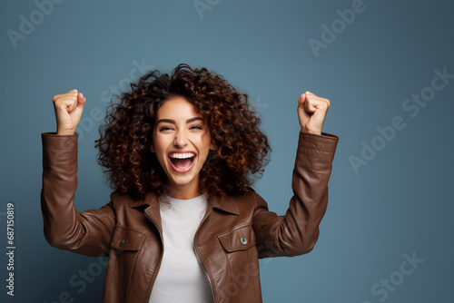 Happy curly  woman in white t-shirt raise hands up isolated on blue background. The concept of winning, good luck or winning photo