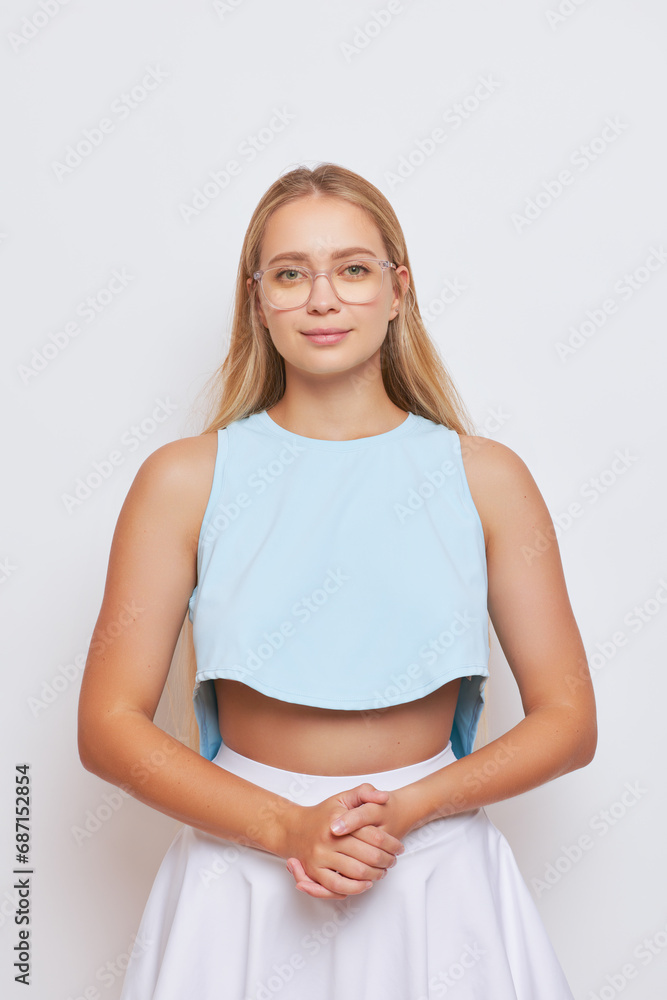 Slender girl with long blond hair in glasses poses with sweet smile on background with her hands folded in front of her, nice day concept, copy space Slender girl with long blond hair in glasses poses with sweet smile on background with her hands folded in front of her, nice day concept, copy space