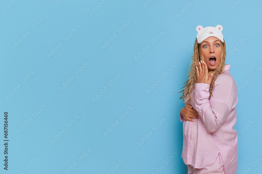 A young expectant mother stands to the side in pink pajamas with sleep mask on head, covering her mouth with hand. good mood concept, copy space A young expectant mother stands to the side in pink pajamas with sleep mask on head, covering her mouth with hand. good mood concept, copy space