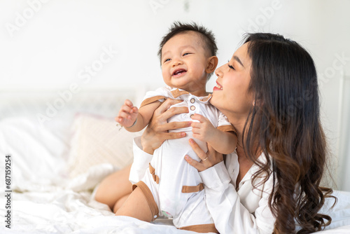 Portrait of enjoy happy love family asian mother playing with adorable little asian baby, newborn, infant.Mom touching care with cute son in a white bedroom.Love of family concept