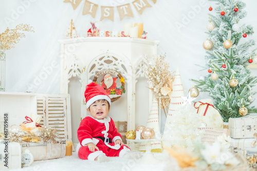 サンタの衣装を着てクリスマスツリーのフォトセットでご機嫌にはしゃぐ1歳の笑顔の赤ちゃん